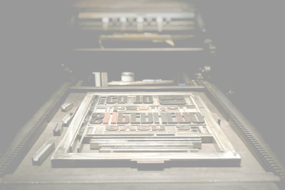 A letterpress chase loaded with wood type on the press bed, photographed in landscape view with the surrounding press machinery in frame
